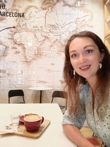 A smiling woman sits at a cafe table with a latte, water, and dessert. Behind her is a world map mural highlighting Barcelona. The ambiance feels cozy.