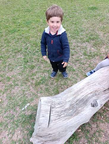 A young boy smiles cheerfully while standing on grass beside a large, weathered log with chalk drawings. Another child’s legs are partially visible on the right.