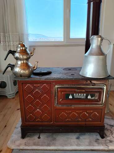 Una estufa de leña antigua se encuentra en una acogedora habitación. Sobre ella hay teteras decorativas y una jarra de metal, con la luz del sol entrando por la ventana detrás.