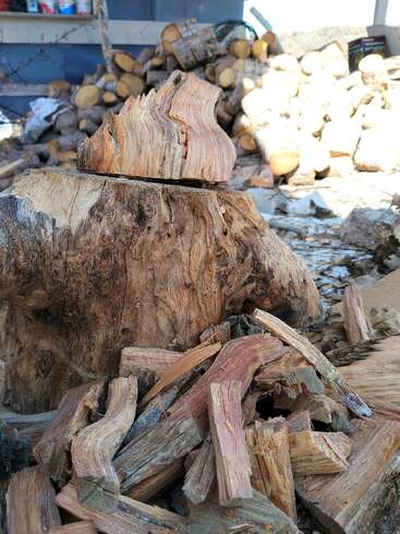 Primer plano de trozos de madera cortada sobre un tocón, con una gran pila de leña al fondo, lo que sugiere un área exterior para cortar y almacenar leña.