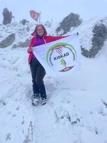 Uma mulher está em pé numa montanha nevada, sorrindo e segurando uma bandeira da KISKAD. Bandeiras turcas são visíveis ao fundo. A neve cobre o terreno rochoso.