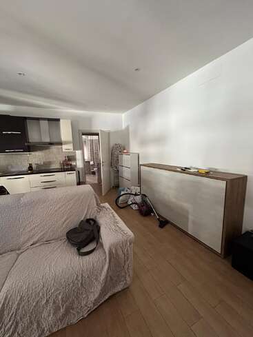 Cette photo montre un salon chaleureux avec un canapé de couleur claire, un coin cuisine, une armoire, des produits d'entretien, des sacs éparpillés et une porte ouverte en arrière-plan.