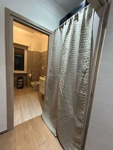 Un couloir mène à une salle de bains aux murs et au sol carrelés dans un style moderne. Des rideaux masquent en partie une autre porte ; une valise noire est posée au-dessus. L'éclairage est chaleureux et accueillant.