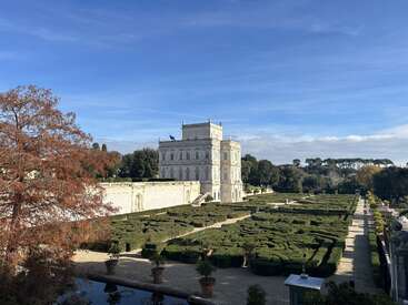 Une imposante villa blanche se dresse majestueusement derrière des jardins à la française soigneusement aménagés, des haies verdoyantes, de grands arbres et un ciel bleu limpide qui s'étend au-dessus du paysage.