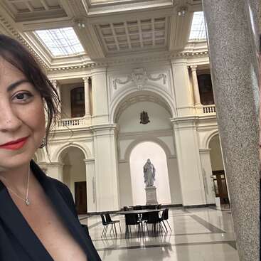 Une femme en tenue noire sourit pour un selfie à l'intérieur d'un grand hall élégant avec de hauts plafonds, des colonnes classiques, une statue centrale et des chaises modernes.