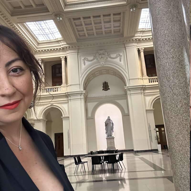 Une femme en tenue noire sourit pour un selfie à l'intérieur d'un grand hall élégant avec de hauts plafonds, des colonnes classiques, une statue centrale et des chaises modernes.