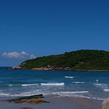 This image shows a serene beach with gentle waves, a sandy shore, rocks, and a lush green hill in the background under a clear blue sky.