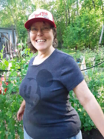 The image depicts a woman standing in a garden, wearing a baseball cap and a dark blue t-shirt with a large black design on the front, surrounded by lush greenery.