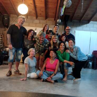 A group of fourteen people, smiling and gathered indoors, pose under warm lighting with wooden beams overhead. Some are sitting on the floor, creating a cozy atmosphere.