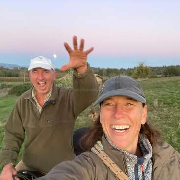 A cheerful man and woman outdoors, both smiling widely. The man waves enthusiastically, wearing a white cap; the woman is in front, wearing a gray cap. Beautiful countryside.