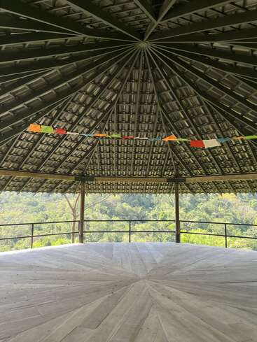 Under a large, circular, open-air wooden pavilion with a geometric roof, colorful prayer flags hang, overlooking lush green forest and serene natural scenery. Peaceful atmosphere.