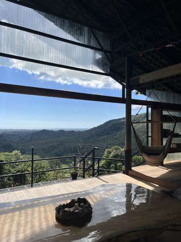 Un acogedor salón al aire libre con hamaca, mesa rústica de madera y vistas panorámicas de la montaña bajo un techo parcialmente cubierto, rodeado de un frondoso bosque verde.