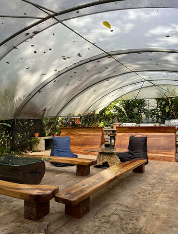 This image shows a cozy outdoor seating area with wooden benches, a glass table, bean bags, and lush plants under a curved, transparent canopy roof.