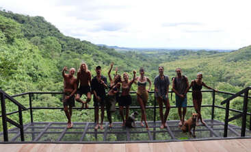 A group of people and two dogs pose on a metal platform, surrounded by lush green forested hills under a cloudy sky, enjoying nature together.