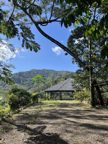 A serene hillside scene with a pavilion surrounded by lush greenery. Blue sky above, mountain views in the background, sunlight and shadows adding peaceful ambiance.