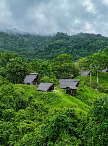 Three small, triangular-roofed cabins sit amidst lush green hills and dense forest. Misty clouds hover over the mountains, creating a serene and peaceful atmosphere.