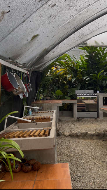 This image shows an outdoor kitchen with a clear roof, concrete sinks, red pots hanging, a stove, lush green plants, gravel floor, and wooden countertops.