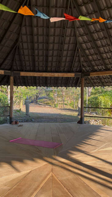 A serene open-air wooden pavilion with a pink yoga mat, colorful flags above, statues on either side, and a lush green nature view outside.