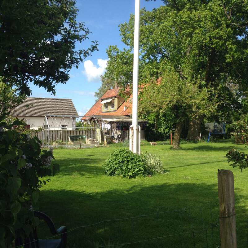 The image depicts a serene backyard scene, featuring a white pole, lush green grass, and a house with a distinctive red roof, surrounded by trees and a fence.