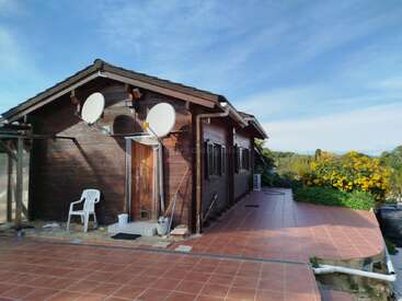 This image shows a wooden house with two satellite dishes, a plastic chair, tiled patio, bright yellow flowers, greenery, and a clear blue sky above.