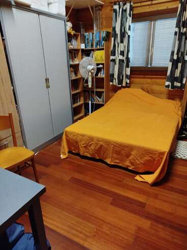 A cozy room with a single bed covered in an orange blanket, a wardrobe, bookshelves, a fan, curtains, a window, and wooden flooring.