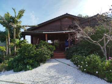 A cozy wooden house with a front porch, surrounded by lush green bushes, blooming flowers, palm trees, and a pebbled pathway under a clear blue sky.