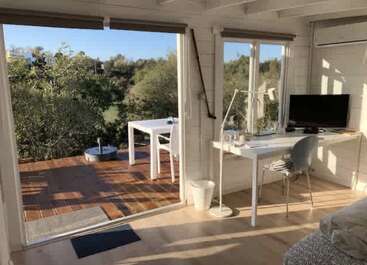 Cette chambre confortable et moderne dispose d'un bureau avec un ordinateur, de grandes fenêtres et d'une porte s'ouvrant sur un patio ensoleillé avec des meubles d'extérieur, entouré de verdure.