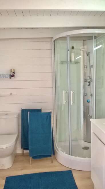 Cette salle de bains comprend une douche moderne en verre, des toilettes blanches, des serviettes bleues, un tapis de bain bleu assorti, un sol en bois et des murs blancs avec une décoration et des étagères minimalistes.