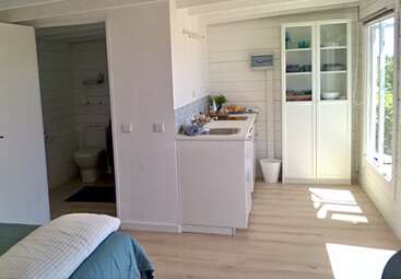 Cette image montre un petit studio lumineux avec une kitchenette, des armoires blanches, une grande fenêtre, une salle de bain visible, des sols en bois, une armoire en verre et un coin lit confortable.