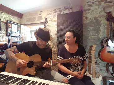 A imagem mostra um homem e uma mulher em uma sala de música, tocando violão e piano, cercados por instrumentos e equipamentos, com um fundo de parede de pedra rústica.