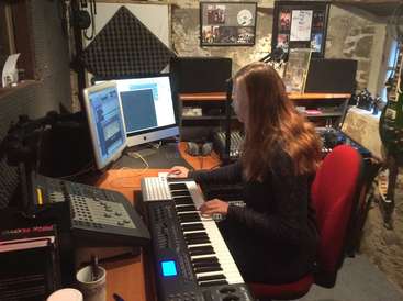 A imagem mostra uma mulher sentada em uma mesa em um estúdio de música, cercada por vários equipamentos, incluindo um teclado e monitores de computador, com uma cadeira vermelha e uma mesa de madeira.