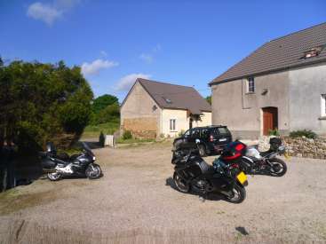 A imagem mostra uma cena rural serena, com uma fileira de motocicletas estacionadas em frente a uma casa rústica e um carro, em um pitoresco céu azul.