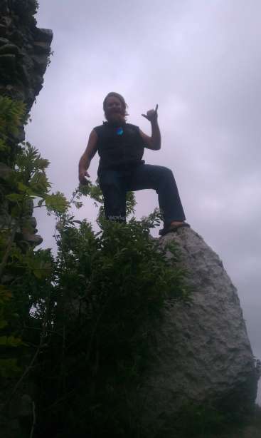 A imagem mostra um homem sentado em uma rocha, fazendo um gesto com a mão, com um céu nublado e vegetação ao fundo. O homem está vestindo uma camisa preta sem mangas e calças.