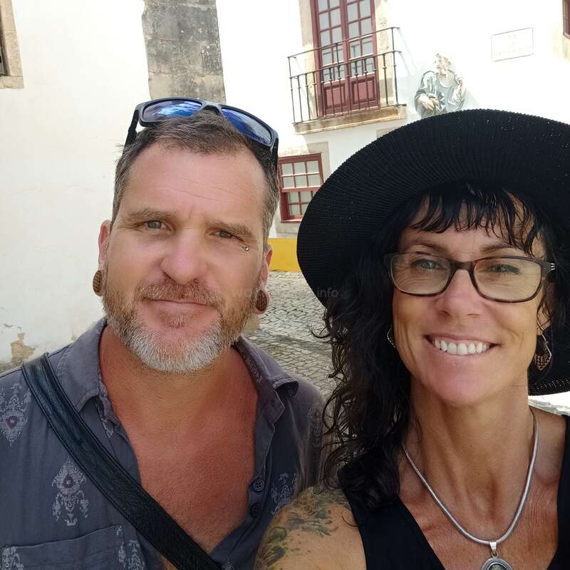 La imagen muestra a un hombre y una mujer posando para un selfie delante de un edificio blanco con una estatua en la pared y un balcón con puertas rojas.