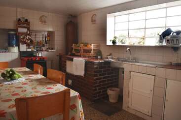 Diese gemütliche Küche hat 'ne Arbeitsplatte aus Backstein, 'nen roten Herd, geflieste Wände, 'ne große Fensterfront, Topfpflanzen, Obstkörbe, Holzstühle und 'nen Tisch mit 'ner Tischdecke mit Blumenmuster drauf.