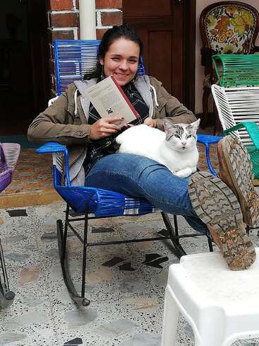 Une femme souriante est assise sur une chaise à bascule bleue, un livre à la main, avec un chat blanc et gris confortablement installé sur ses genoux, profitant d'un moment de tranquillité.