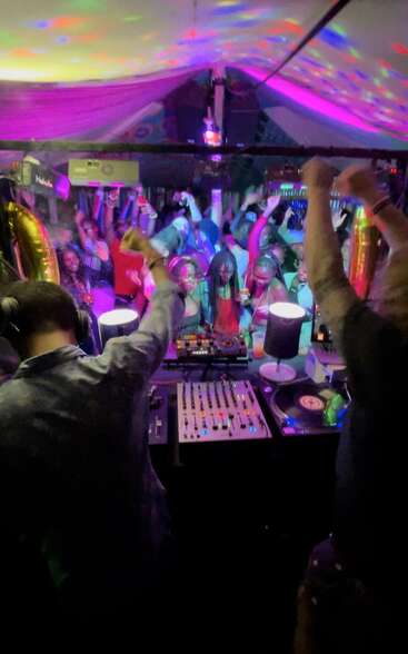 A vibrant nightclub scene with a DJ spinning tracks, colorful lights, dancing crowd with raised arms, joyful energy, and people celebrating together under a glowing canopy.