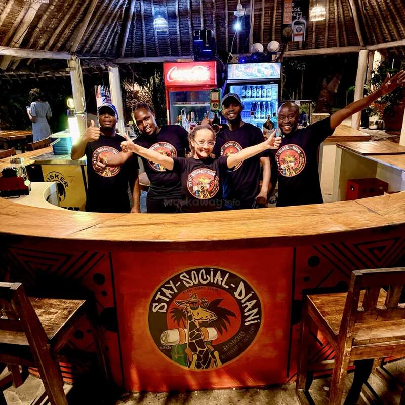 Five smiling staff members stand behind a wooden bar with "Stay Social Diani" logo, wearing matching shirts, posing cheerfully in a vibrant, tropical, open-air setting.
