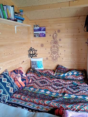 Una acogedora habitación de madera con ropa de cama de estampados coloridos, cojines decorativos, cuadros en las paredes, una estantería con libros y fotografías, y un ambiente tranquilo y bohemio que invita al descanso y al confort.