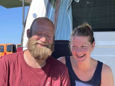 Un homme barbu et une femme souriante sont assis côte à côte devant un camping-car. La journée ensoleillée et leurs visages détendus et joyeux laissent penser qu'ils profitent d'un moment agréable en camping.