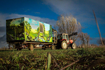Un tracteur rouge tire une remorque ornée d'une fresque colorée représentant un paysage. La scène se déroule dans un champ verdoyant, sous un ciel spectaculaire.