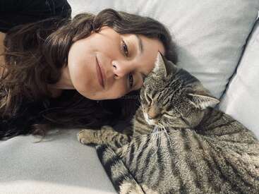 Eine Frau liegt auf einer hellen Couch und ruht ihren Kopf neben einer entspannten getigerten Katze. Beide wirken friedlich und genießen einen gemütlichen und liebevollen Moment miteinander.