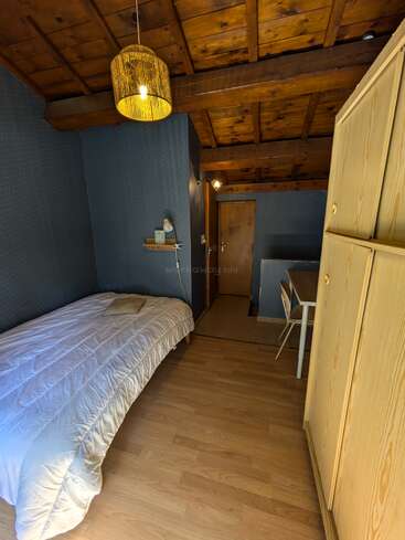 Dieses gemütliche Zimmer verfügt über Holzböden und -decken, ein Einzelbett mit weißem Bettzeug, einen Kleiderschrank, einen Schreibtisch mit Stuhl, ein Regal und eine warme Hängelampe.