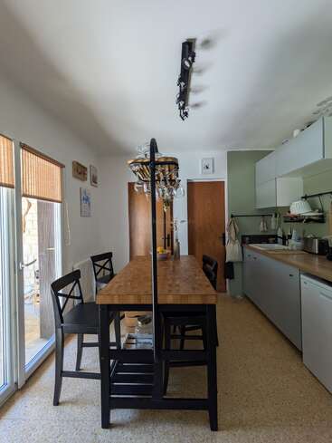A modern kitchen with wooden countertops, black chairs, a wooden table, hanging glasses, light fixtures, wall art, appliances, and large windows with bamboo blinds. Cozy atmosphere.