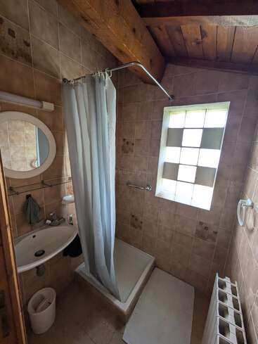 This image shows a small rustic bathroom with brown tiled walls, a wooden ceiling, a simple shower with a curtain, a window, sink, mirror, and radiator.