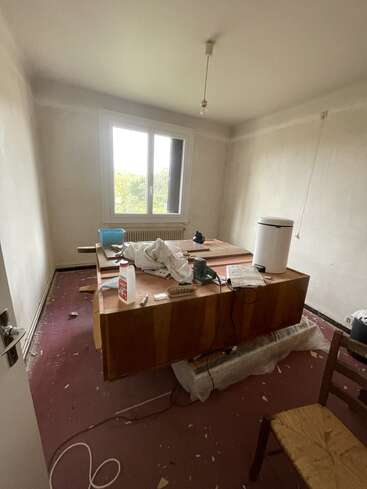 A small, unfinished room with bare walls and a single window. The floor is messy, and a large desk is cluttered with renovation supplies and tools.