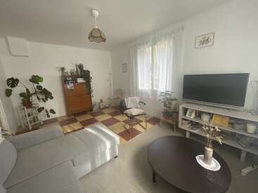 The image shows a cozy living room with a sectional sofa, TV, coffee table, indoor plants, checkered floor tiles, white walls, and soft natural light.