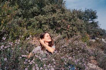 A woman sits among blooming wildflowers, smiling and enjoying the sunshine. She looks peaceful and content, surrounded by lush green bushes and a clear blue sky.