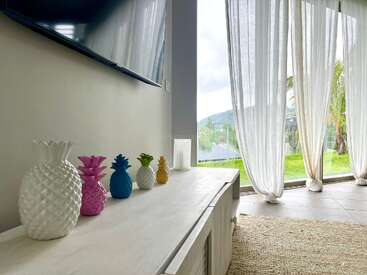 Un luminoso y moderno salón con cortinas blancas, coloridos adornos de piñas sobre un mueble blanco, un gran televisor y vistas a la vegetación exterior.