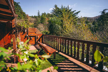 Un balcon en bois avec un banc surplombe des arbres et des collines verdoyantes sous un ciel bleu clair, créant une retraite paisible et invitante en plein air.